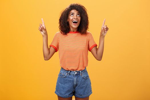 Fascinated impressed and amused good-looking charming African-American woman with afro hairstyle in trendy t-shirt and denim shorts pointing and looking up with interested look over orange wall. Lifestyle.