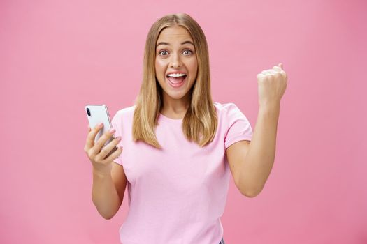Girl beat own record in smartphone game raising clenched fist in cheer and triumph holding cellphone, smiling excited and happy at camera celebrating victory with joyful gesture over pink background. Technology and advertising concept