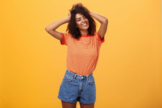 Girl feeling awesome making cool hairstyle posing near mirror feeling glad to have date tonight holding hands on curly hair smiling joyfully and tender standing against orange background. Lifestyle.