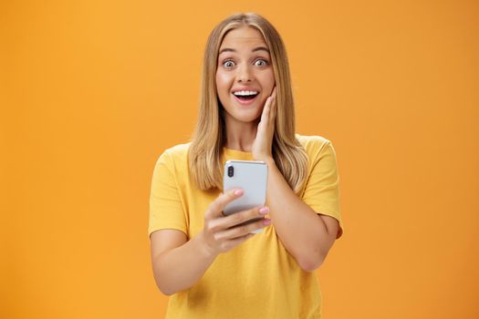 Portrait of surprised and impressed woman reacting to awesome app in smartphone touching cheek from amazement and joy smiling broadly at camera holding cellphone in hand over orange wall. Technology and emotions concept