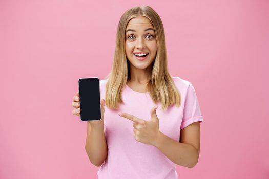 Cheerful attractive and pleasant woman promoting cool app or smartphone holding cellphone and pointing at device screen smiling amused and impressed standing over pink background. Technology concept