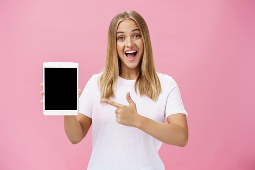 Woman giving recommendation buy digital tablet pointing at device screen with index finger smiling enthusiastic and pleased telling own opinion about gadget perfomance posing delighted over pink wall. Lifestyle.