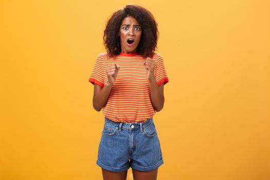 Woman about to panic hearing shocking worried news. Portrait of intense nervous and anxious dark-skinned adult girl with afro hairstyle clenching fists opening mouth looking concerned over orange wall.