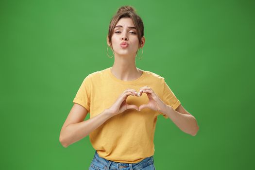 Sensual charming woman in yellow t-shirt and combed hairstyle in yellow t-shirt showing heart gesture against chest folding lips to give kiss or mwah tender and loving over green background. Lifestyle.