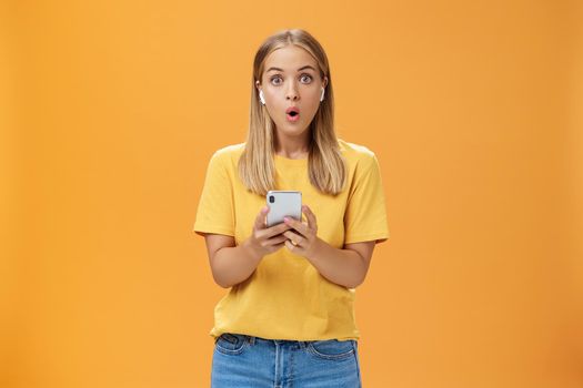 Woman surprised how awesome sounds new wireless earbuds holding smartphone wearing earphones folding lips in wow and amazement listening music impressed by good product over orange background. Copy space