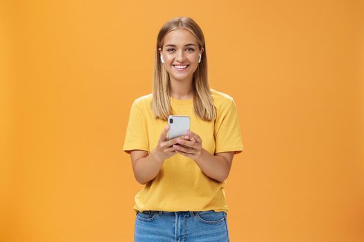 Portrait of friendly charismatic and outgoing good-looking young female in yellow t-shirt holding smartphone wearing wireless earphones enjoying listening music on way to fitness gym. Technology and lifestyle concept