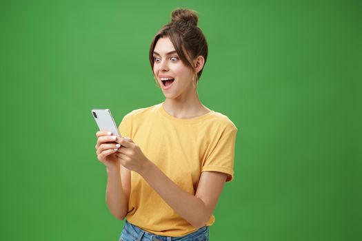 Woman reading surprising satisfying message in smartphone opening mouth from excitement, smiling amazed looking astonished at cellphone screen posing against green background in casual yellow t-shirt. Technology concept