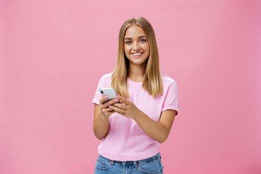 Woman calling taxy via smartphone asking friend address smiling cheerfully at camera holding phone with both hands over chest getting in touch with clients via messages standing over pink wall. Technology concept