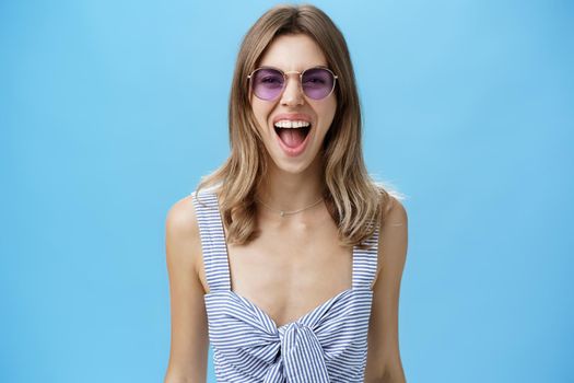 Expressive and careree woman being wild ad free yelling out loud at camera wearing awesome stylish sunglasses and striped top letting out emotions being open-minded and rebellious over blue wall. Lifestyle.