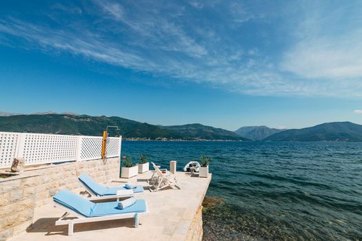 white wooden sunbeds on the pier, by the sea. In the Gulf of Kotor, in Montenegro. Azure blue water and pebble bottom, mountain views and blue sky with white clouds. High quality photo