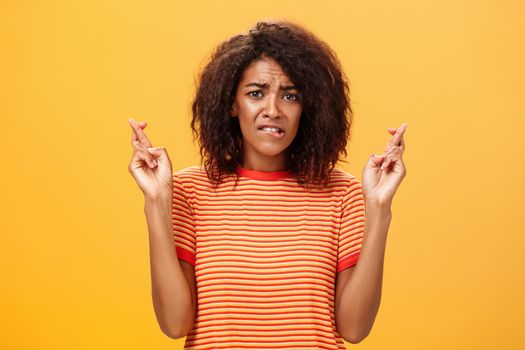 Waist-up shot of concerned troubled nervous african american woman with curly hairstyle biting lower lip anxiously frowning crossing fingers for good luck hopefully praying for dream come true. Lifestyle.