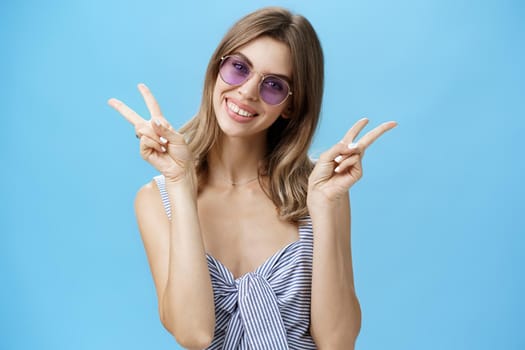 Waist-up shot of cute self-assured feminine girlfriend in trendy top showing peace gestures near shoulders tilting head amused smiling broadly showing off gapped teeth posing over blue wall. Lifestyle.