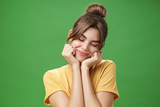 Tender sensual timid european girl in yellow t-shirt leaning head on shoulder touching cheeks with hands closing eyes and smiling with soft grin feeling nostalgic and romantic over green background. Lifestyle.