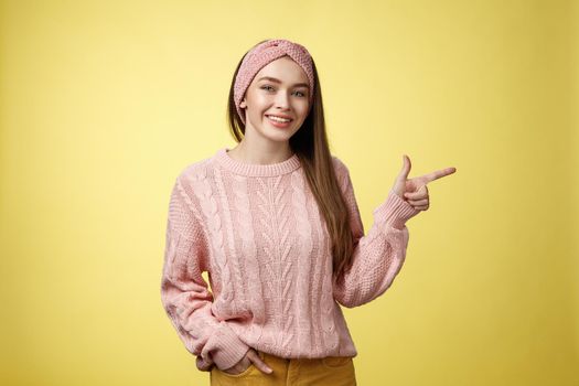 Lifestyle, people, pointing concept. Attractive young girl with long straight hair wearing sweater pointing aside using fore finger, indicating copy space smiling cheerful, positive over yellow wall.