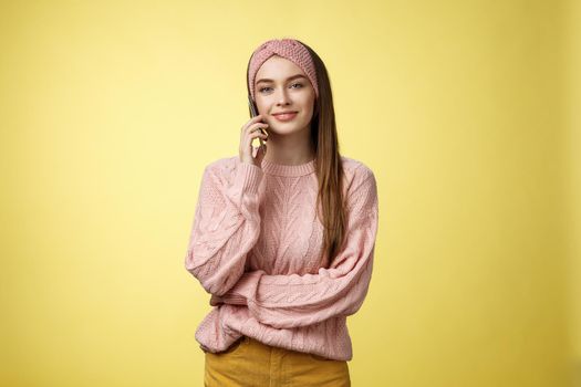 Stylish young girlfriend having pleasant conversation via phone holding smartphone near ear talking, making reservation in restaurant, ordering food delivery, smiling, posing against yellow wall.