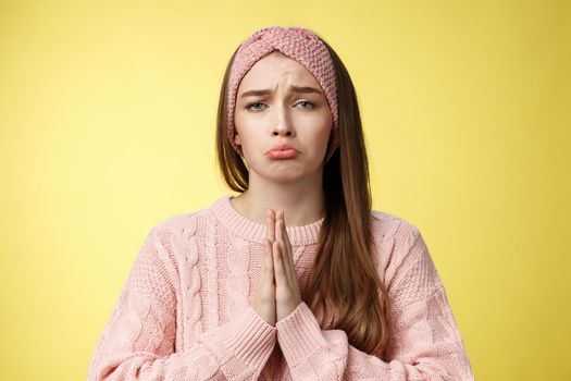 Clingy sad whining girlfriend bagging buy her dress grimacing pouting and frowning upset, pressing palms together in pray, asking favour, apologizing, praising friend to help over yellow background.