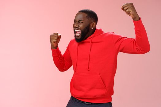 Extremely happy bearded young 25s black guy triumphing happily yelling yeah raising clenched fists celebrating success winning bet lottery standing happily pink background cheer receive great news.