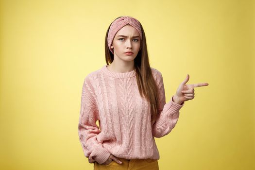 Pissed puzzled young attractive arrogant moody girlfriend in sweater, headband looking irritated, intense frowning pointing finger at copy space, directing person sits on her place, annoyed.