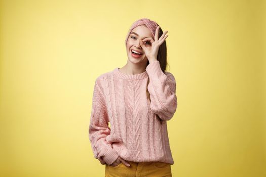 Cheerful girl saying ok. Attractive stylish shoolgirl in knitted sweater relaxed feeling upbeat, sharing happiness and joy, showing okay sign on eye smiling optimistic, entertained over yellow wall.