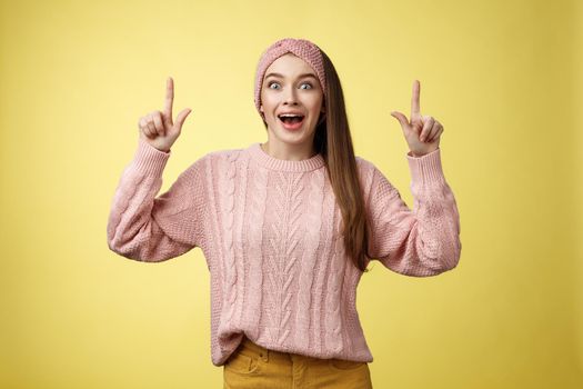 Ecstatic amused positive young 20s woman wearing casual sweater, headband dropping jaw amazed looking astonished, surprised pointing up excited, upbeat of seeing incredible promo over yellow wall.