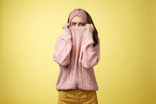 Sad silly, gloomy cute young girl hiding face in sweater collar pulling clothes on nose frowning displeased, unhappy, looking indignant and reluctant standing disappointed over yellow background.