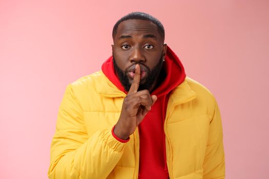 Waist-up serious-looking african-american bearded man in yellow jacket red hoodie bend camera asking keep quiet say shhh show shush gesture index finger on lips, confidential information taboo.