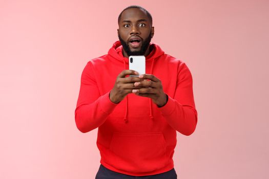 Impressed focused handsome african american bearded guy looking intrigued holding smartphone recording video capturing awesome fight, widen eyes curious surprised, standing pink background.