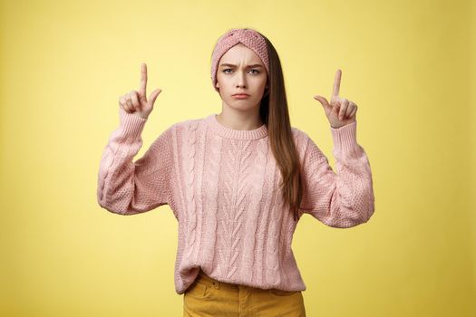 Moody disappointed and upset unamused cute young european girl in knitted warm sweater headband expressing dislike and disagreement pointing up, sulking frowning displeased unhappy with bad choice.