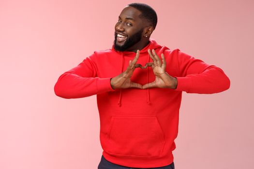 Someone love ya. Portrait enthusiastic creative cute black boyfriend wearing red hoodie show heart sign smiling broadly confessing love sympathy look passionate, express romance pink background.