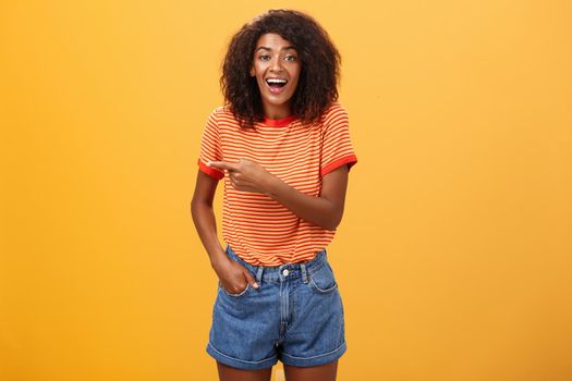 Woman laughing over funny puppy. Portrait of amused and entertained attractive stylish african-american female in casual denim shorts pointing left talking about awesome copy space over orange wall.