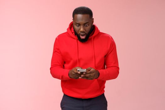 Amused focused impressed african-american bearded guy in red hoodie drop jaw widen eyes look amazed smartphone display winning hard game level answering exciting message, standing pink background.