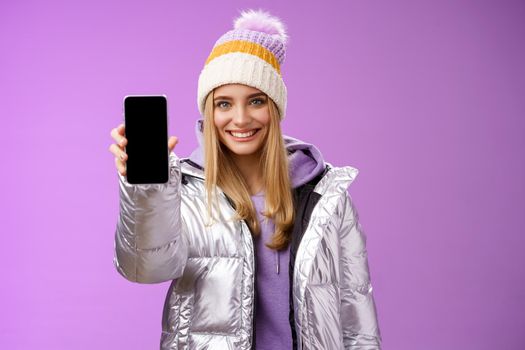 Lifestyle. Friendly cheerful confident blond girl in silver stylish winter jacket hat extend arm showing smartphone display advertising awesome new device app smiling self-assured recommend use mobile phone.