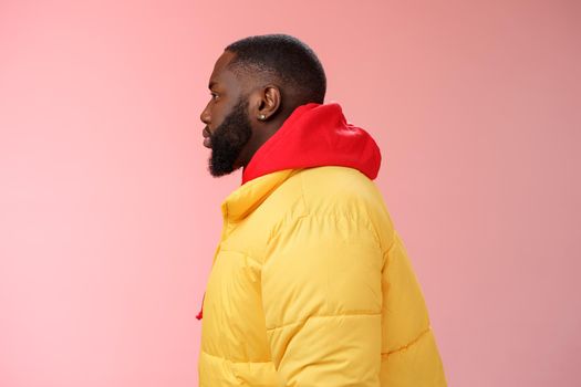 Profile studio shot bearded young 25s african guy in yellow jacket red hoodie look left normal unbothered relaxed expression standing queue order fastfood pink background, waiting take-away.