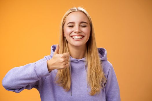 Amused joyful optimistic young excited blond european female model in hoodie smiling broadly close eyes having fun show approval gesture thumbs-up like awesome trip vacation idea, orange background.