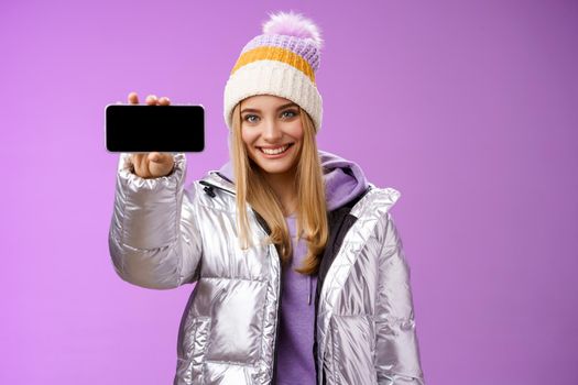 Confident friendly good-looking blond girl in outdoor silver glittering jacket hat hold smartphone horizontal showing mobile phone display assertive smile recommend use app, purple background.