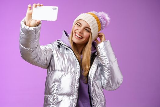 Joyful carefree charming blond girl having fun wanna post pictures from ski resort vacation taking seflie holding smartphone mimicking show tongue winking look mobile display, purple background.