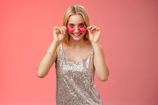Lucky attractive stylish european blond female having fun friends nightclub wearing silver glittering dress sunglasses look amused curious peek camera intrigued, standing red background.