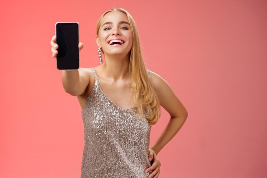 Proud joyful charming cheerful blond european woman in stylish silver shiny dress hold hand waist confident extend arm showing smartphone display present awesome new app device, red background.