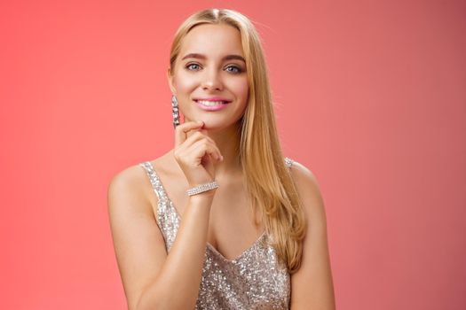 Waist-up shot elegant good-looking wealthy young blond woman in brilliant accessorize silver glittering stylish dress touch chin have clever interesting idea smiling curiously, red background.