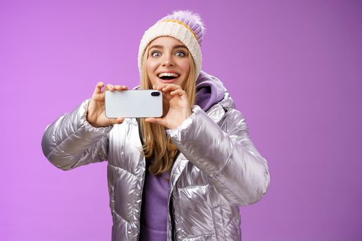 Amused fascinated female in silver jacket head smiling astonished excited look forward holding smartphone recording video taking shots famous person mobile phone camera, purple background.