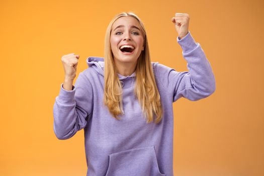 Happy cheerful young supportive woman cheering sister win smiling dedicated fun hope favorite team score goal raising clenched fists yelling happily celebrating triumphing victory, orange background.