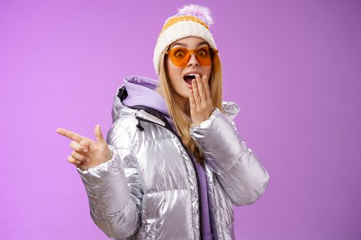 Amazed speechless surprised attractive stylish blond girlfriend in sunglasses, silver glittering jacket hat drop jaw impressed widen eyes shocked pointing left astonished, purple background.
