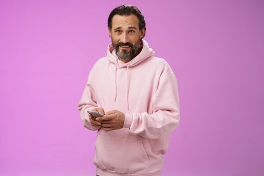 Stylish handsome adult bearded guy 40s in trendy pink hoodie holding smartphone texting look camera unbothered cool confident distracted playing awesome new app game, purple background.
