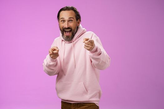 Gotcha. Portrait carefree funny amusing adult energized bearded man jumping pranking friend having fun laughing happily pointing camera index fingers greeting picking you, purple background.