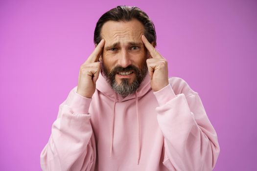 Distressed pressured intense worried mature caucasian man bearded grey hair frowning grimacing thinking hold hands temples bothered cannot make up plan idea, have crisis, purple background.