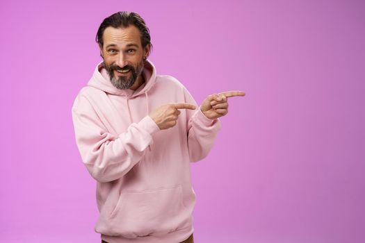 Sassy confident good-looking macho man 50s in pink hoodie smiling broadly inviting join pointing right showing interesting amusing place hang out welcoming take look, standing purple background.