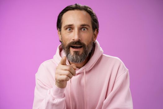 Fascinated amused european bearded mature guy widen eyes shocked open mouth recognize person pointing camera index finger surprised see familiar famous star, standing purple background.