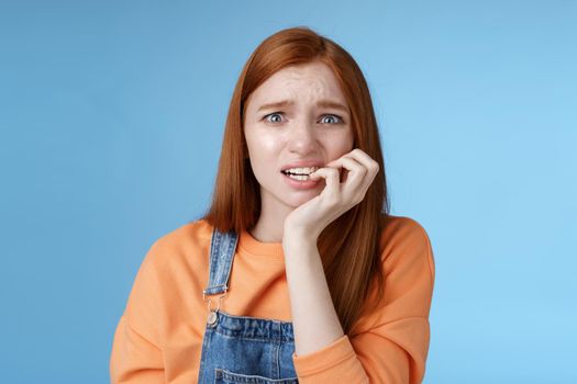 Intense worried scared unsure young redhead panicking silly girl frowning look upset anxious biting fingernails emotional frightened fired standing blue background emotional terrified. Copy space