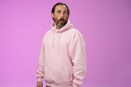 Impressed speechless shocked adult bearded man grey hair in cool pink hoodie folding lips wow popping eyes camera surprised astonished intrigued, standing curious amazed purple background.