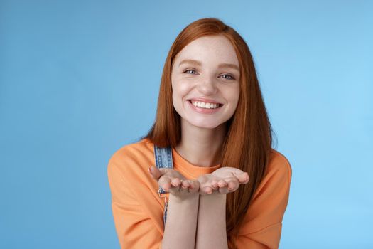 Cute tender kind young ginger girl giving all love you, hold something palms showing camera smiling delighted introduce present grinning romantic gesture send air kisses, blue background.